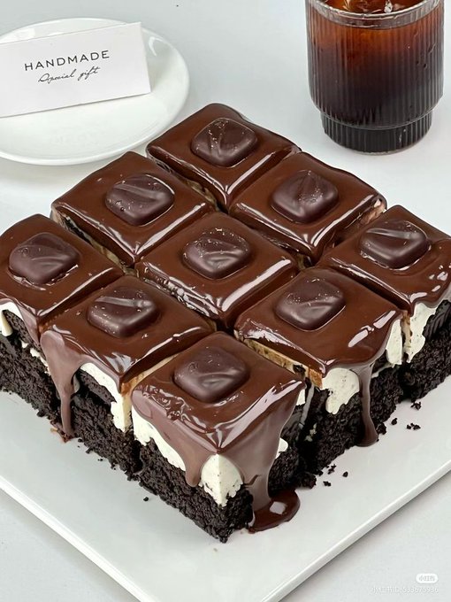 A square chocolate cake cut into nine equal pieces, each topped with a smaller square of chocolate. The cake has layers of dark chocolate, white cream, and a brownie base, with chocolate drizzle flowing down the sides. A white plate holds the cake, and a glass of iced coffee is visible in the background. A card labeled "HANDMADE Special Gift" is placed on a smaller plate beside the cake. An Instagram watermark is present.