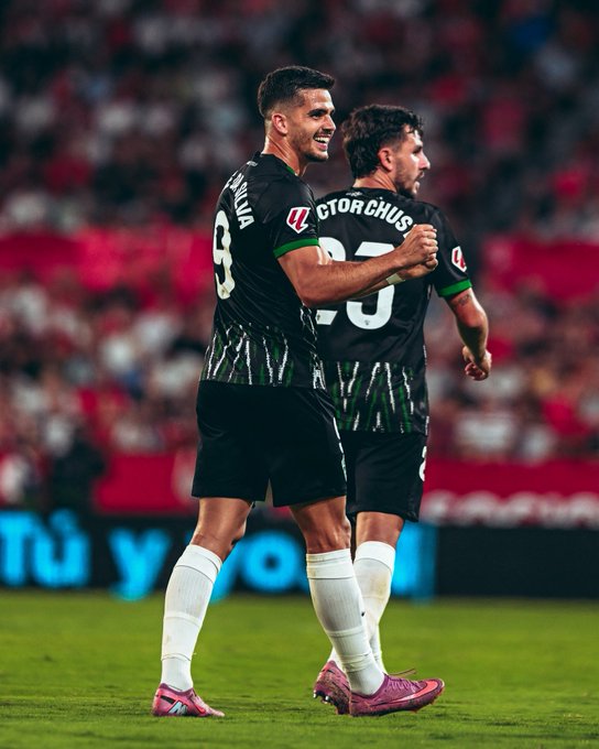 Two soccer players wearing black and green Elche CF jerseys with the numbers 9 and 20 on the shorts. The player with number 9 has dark hair and is pointing forward, wearing pink cleats and white socks. The player with number 20, named Víctor Chust, has dark hair and is smiling, walking beside the other player on a grassy field. A large crowd is visible in the background, and red stadium seating is seen.