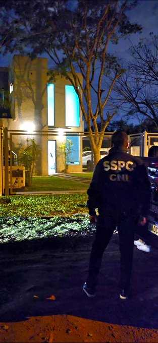 A person wearing a dark jacket with "SSPC" and "CNI" text on the back stands outdoors at night, facing a modern house with large vertical windows illuminated in green. A tree and grass are visible in the yard, and a vehicle is partially seen near the house. The scene is well-lit by artificial lighting.