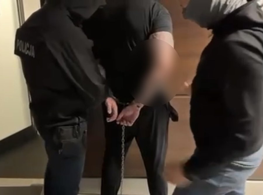 Two police officers in dark uniforms with "POLICJA" on the back stand on either side of a handcuffed man in a black shirt and pants. The man is restrained with metal handcuffs and a chain, wearing a blurred face for anonymity. Another individual in a dark jacket and light pants stands nearby, also with a blurred face.