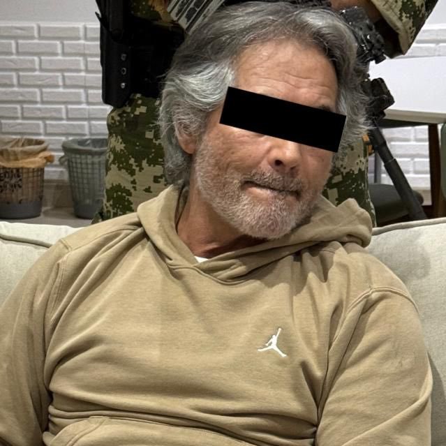 Hernán Bermúdez Requena sentado en un sofá de color claro, con una sudadera beige con un pequeño logo blanco. Se le ven las canas y tiene barba. Una persona con atuendo militar de camuflaje está detrás de él, parcialmente visible.
