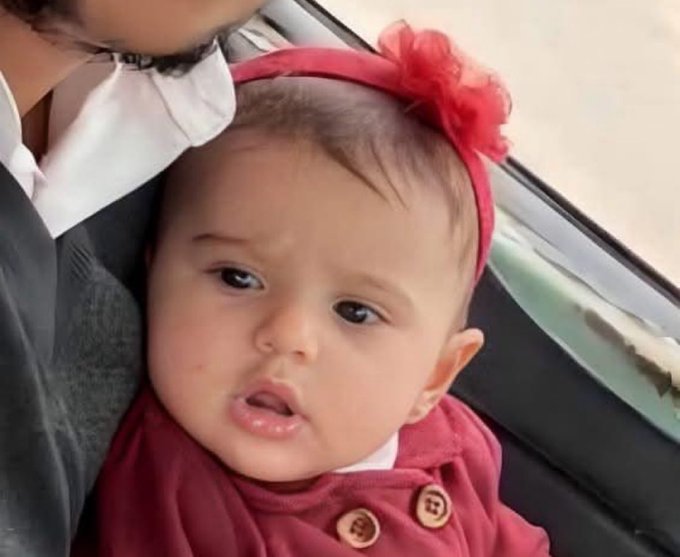 Laila Khatib, a baby wearing a red outfit and a red headband with a bow, is held by an adult in a car. The baby has light skin, dark hair, and is dressed in a red buttoned top.