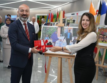 A group of people in formal attire holding red ribbons, cutting a ceremonial ribbon in a modern indoor space with flags from various countries in the background. A woman in a headscarf and light-colored coat stands among them, holding a booklet. Easels display large photographs of families, including one of a woman and child in colorful clothing. Another image shows a man in a suit presenting a red gift box to a woman in a black dress near a photograph of people together. A third image depicts three people examining photographs on easels, with a Turkish flag visible. Text on a sign reads "FOTOGRAF TARIHIMASİ."