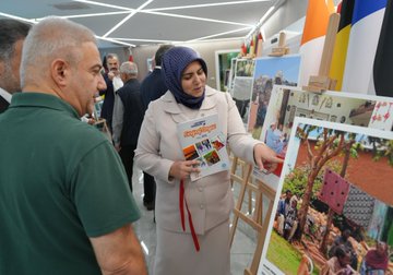 A group of people in formal attire holding red ribbons, cutting a ceremonial ribbon in a modern indoor space with flags from various countries in the background. A woman in a headscarf and light-colored coat stands among them, holding a booklet. Easels display large photographs of families, including one of a woman and child in colorful clothing. Another image shows a man in a suit presenting a red gift box to a woman in a black dress near a photograph of people together. A third image depicts three people examining photographs on easels, with a Turkish flag visible. Text on a sign reads "FOTOGRAF TARIHIMASİ."