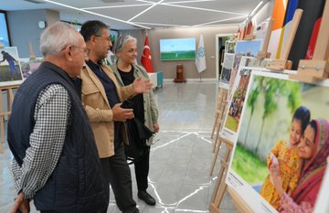 A group of people in formal attire holding red ribbons, cutting a ceremonial ribbon in a modern indoor space with flags from various countries in the background. A woman in a headscarf and light-colored coat stands among them, holding a booklet. Easels display large photographs of families, including one of a woman and child in colorful clothing. Another image shows a man in a suit presenting a red gift box to a woman in a black dress near a photograph of people together. A third image depicts three people examining photographs on easels, with a Turkish flag visible. Text on a sign reads "FOTOGRAF TARIHIMASİ."