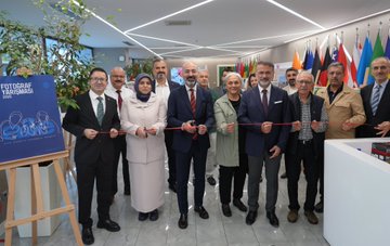 A group of people in formal attire holding red ribbons, cutting a ceremonial ribbon in a modern indoor space with flags from various countries in the background. A woman in a headscarf and light-colored coat stands among them, holding a booklet. Easels display large photographs of families, including one of a woman and child in colorful clothing. Another image shows a man in a suit presenting a red gift box to a woman in a black dress near a photograph of people together. A third image depicts three people examining photographs on easels, with a Turkish flag visible. Text on a sign reads "FOTOGRAF TARIHIMASİ."