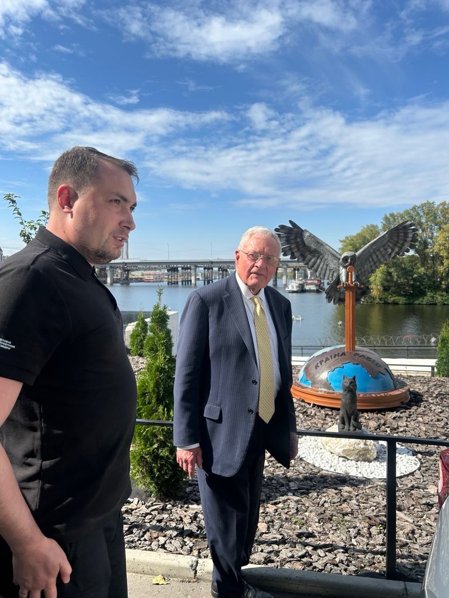 Keith Kellogg in a suit with a yellow tie stands outdoors near a statue of an eagle perched on a globe with a cat figure. A man in a black shirt and pants is beside him. Trees, a river, and a bridge are visible in the background under a partly cloudy sky.