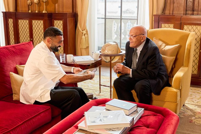 Mohamed Salah sitting on a red couch, wearing a white shirt and black pants, holding a small object. An older man in a suit and tie sits on a yellow armchair, holding a book. A globe and books are on a small table between them, with red and gold cushions visible. Wooden paneling and large windows with curtains form the background.