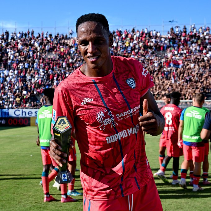 Yerry Mina wearing a red Cagliari soccer jersey with "Sardegna" and a logo, holding a trophy in his left hand, giving a thumbs-up with his right hand. Other players in similar red and green jerseys are visible in the background on a grassy field. A large crowd fills the stands under a clear blue sky. Text on a scoreboard reads "Ches 0".