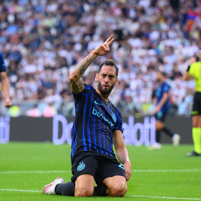 A soccer player wearing an Inter Milan jersey with black and blue vertical stripes and the "betsson" logo, kneeling on a grassy field. He has tattoos on his arms and is making a peace sign with his right hand. A large crowd of spectators is visible in the background, along with a referee in a yellow shirt.