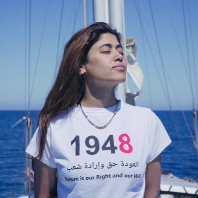 Rima Hassan standing on a boat with the sea and sky visible in the background. She wears a white T-shirt with the text "1948" in large red numbers, Arabic text, and the English phrase "Return is Our Right and Our Will" printed below. A necklace is visible around her neck, and the boat’s mast and rigging are seen behind her.
