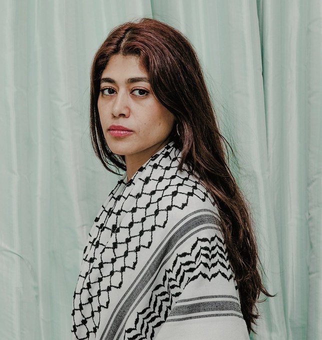 Rima Hassan with long brown hair, wearing a patterned black and white keffiyeh draped over her shoulders, standing against a light green curtain background.