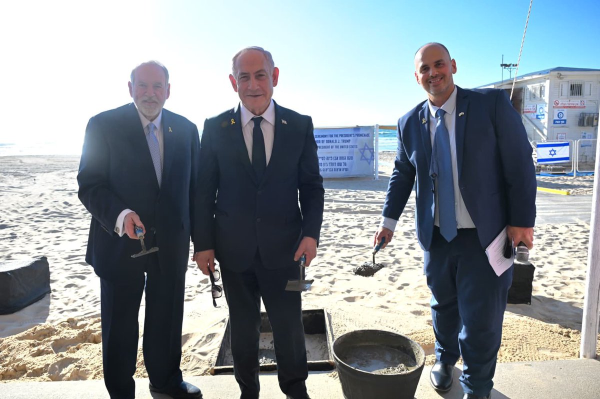 Benjamin Netanyahu, Mike Huckabee, and Tzvi Brot standing on a beach. They are dressed in suits and ties, holding shovels. A concrete bucket and sand are visible. Blue signs with Hebrew text are in the background.