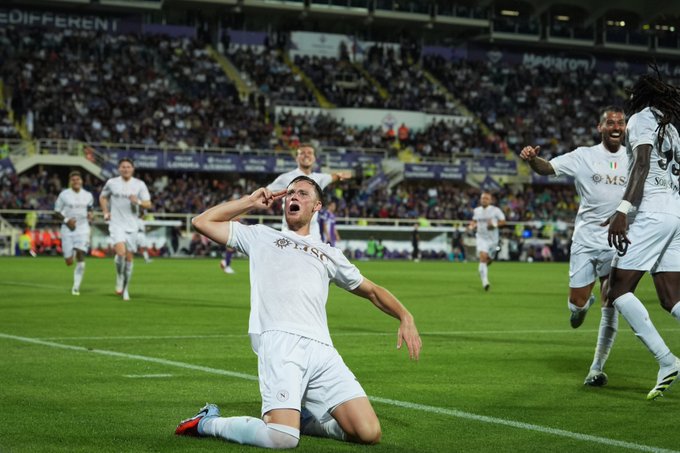 Sam Beukema kneeling on a soccer field, wearing a white uniform with blue accents, celebrating with arms raised. Other players in similar white uniforms are running and celebrating on the grass. A large stadium with a crowd is visible in the background.