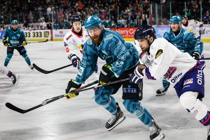 Ice hockey players in action on an ice rink. Belfast Giants players wear teal and white uniforms with intricate patterns, while Dundee Stars players wear white and navy uniforms with "Elite" text. Players skate and handle hockey sticks, engaging in a physical play near the boards, with a crowd visible in the background. Advertisements for "Harry Corry" and phone numbers are on the rink boards.