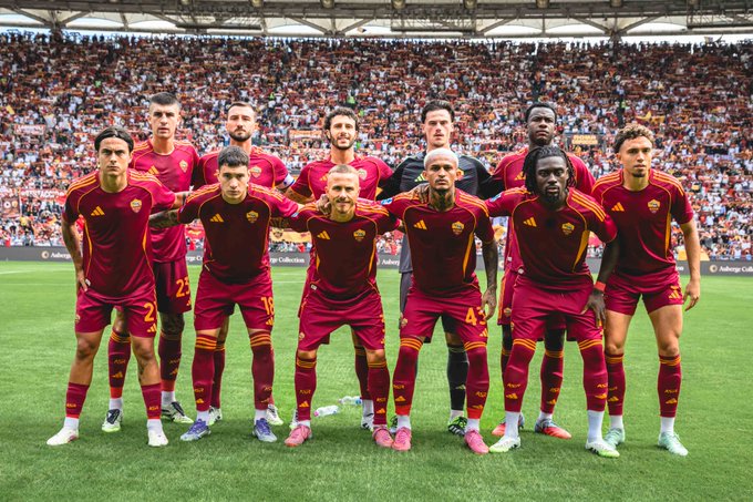 A group of AS Roma soccer players in maroon uniforms standing on a grassy field. They are arranged in two rows, with some players standing and others kneeling, arms linked. A large stadium crowd is visible in the background.