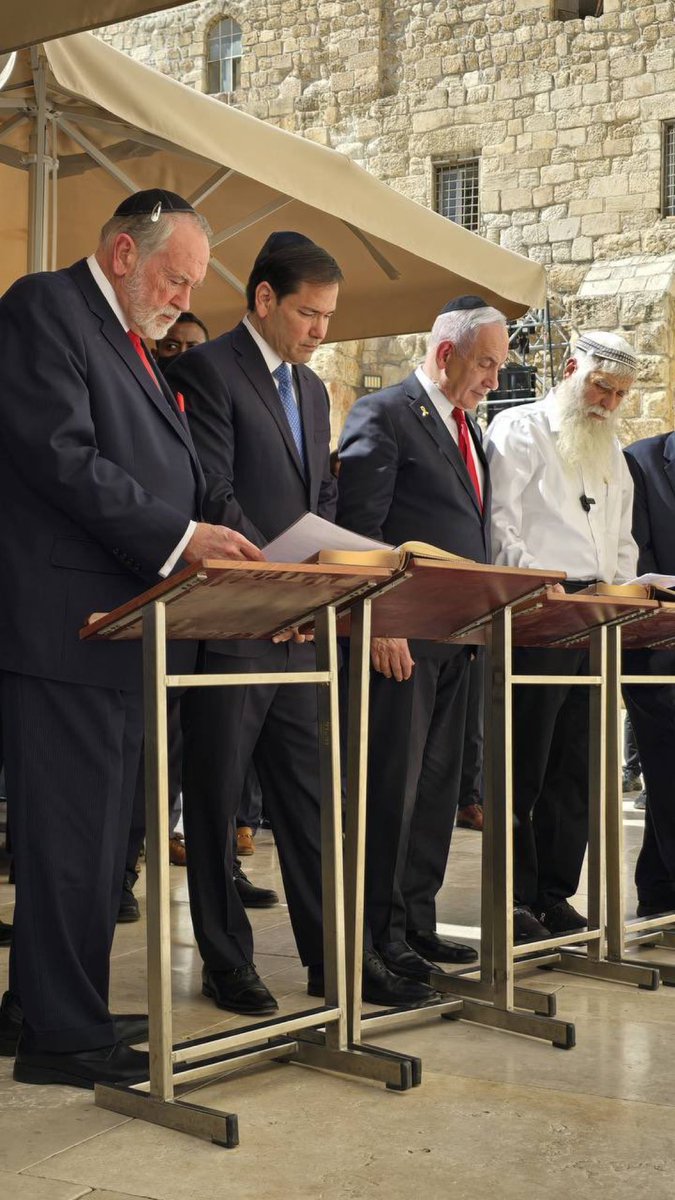 Four men in suits and formal attire standing at podiums with open books. The setting includes a stone building and a beige canopy in the background.