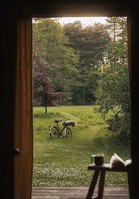 A view through an open doorway showing a lush green meadow with trees in the background. A bicycle is parked on the grass. A wooden table with a cup and books is visible on the left side of the doorway.