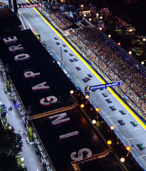 Una vista nocturna de la pista de carreras del Gran Premio de Singapur, con múltiples autos de Fórmula 1 compitiendo en el circuito. La pista está iluminada con luces brillantes y las tribunas llenas de espectadores se alinean a los lados. Grandes letreros que muestran "SINGAPORE" son visibles en los edificios adyacentes a la pista.