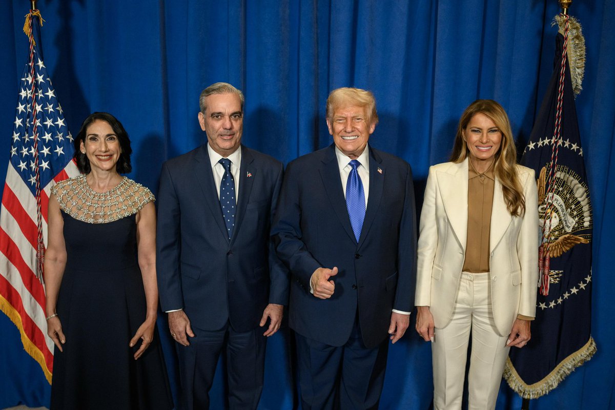 Luis Abinader, Raquel Abinader, Donald Trump, and Melania Trump standing side by side. They are dressed in formal attire, with Luis Abinader and Donald Trump in suits and ties, Raquel Abinader in a sleeveless dress, and Melania Trump in a light-colored blazer and pants. Two flags, one American and one with an eagle emblem, are visible on either side of the group, with a blue curtain backdrop.