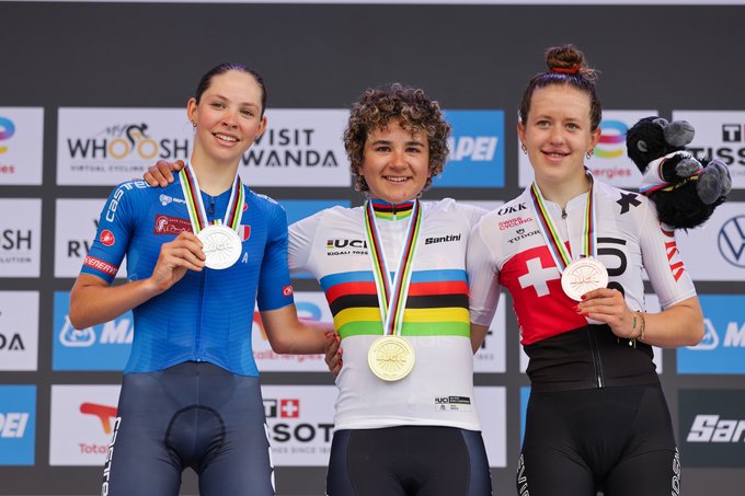 Paula Ostiz, Chantal Pegolo, and Anja Grossmann stand on a podium wearing cycling uniforms. Paula Ostiz in the center wears a white jersey with rainbow stripes and a gold medal. Chantal Pegolo on the left wears a blue jersey with a silver medal. Anja Grossmann on the right wears a white and red jersey with a bronze medal. Sponsors and logos are visible on the backdrop.