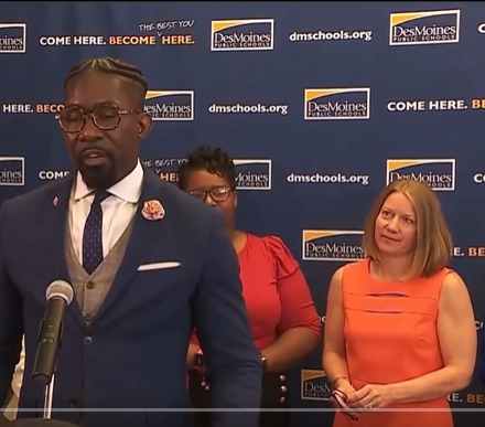 A man in a blue suit, vest, and tie with glasses and braids stands at a podium speaking into a microphone. A woman in an orange dress stands beside him, holding glasses. A backdrop displays repeated "Des Moines" text and "dmschools.org" logos with the slogan "COME HERE. BECOME HERE."