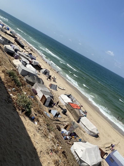 A sandy beach along the Gaza shore with numerous tents and makeshift shelters scattered across the sand. The ocean waves are visible, extending to the horizon under a clear blue sky. People are seen near the tents and along the shoreline.