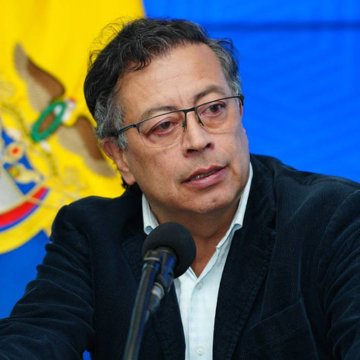 A man with dark hair and glasses wearing a dark jacket over a white shirt, speaking into a microphone. A yellow and blue flag with a coat of arms is partially visible in the background. The Israeli flag with blue stripes and a Star of David is shown separately.