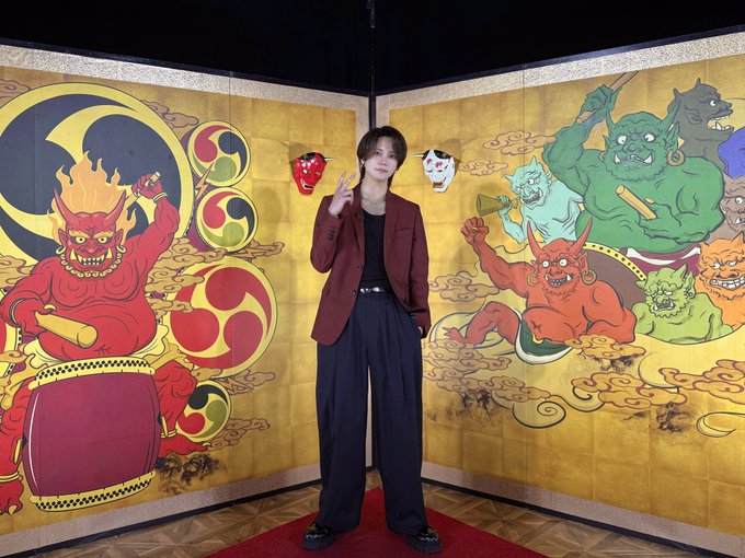 Kyosuke Fujimaki stands in front of vibrant painted panels featuring oni demons and traditional Japanese motifs, including red and green creatures with horns, clouds, and circular patterns. He wears a dark blazer, black shirt, and wide-leg pants, holding a red object in one hand and making a peace sign with the other. Two masks, one red and one white, hang on the panels behind him.