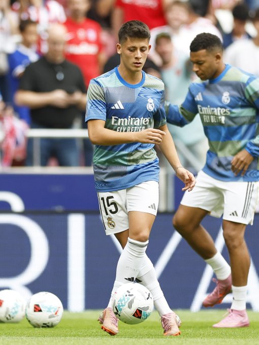 Arda Guler wearing a blue and white Real Madrid jersey with the number 15, dribbling a soccer ball on a grassy field. Another player in a matching Real Madrid uniform is visible in the background.