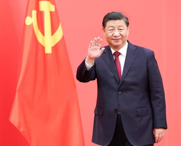 Xi Jinping waving, wearing a dark suit and red tie, standing in front of a Chinese flag with a yellow hammer and sickle. A Palestinian flag waving against a dramatic orange and cloudy sky.