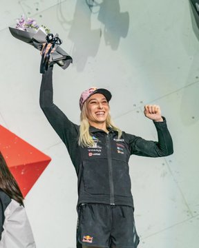 Janja Garnbret stands on a podium, raising both arms in celebration, holding a bouquet of flowers in her left hand. She wears a black athletic jacket and shorts with various sponsor logos, including Red Bull, and a cap. A green backdrop displays text and logos, including "IFSC CLIMBING & PARA CLIMBING WORLD CHAMPIONSHIPS SEOUL 2025" and sponsor names like EP and Kalas.