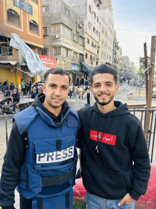 Two men standing on a busy urban street. One wears a blue press vest with "PRESS" text over a black hoodie. The other wears a black hoodie with red Arabic text. Buildings, people, and street activity are visible in the background.