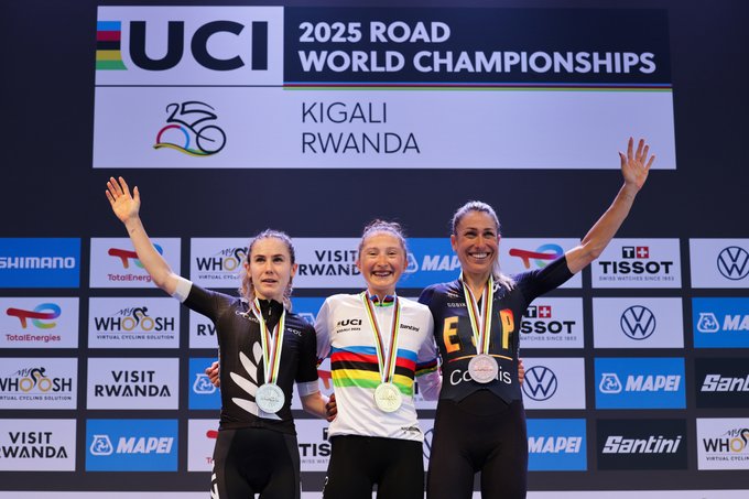 Magdeleine Vallieres, Niamh Fisher-Black, and Mavi García standing on a podium. Magdeleine Vallieres wears a white and black cycling outfit with a gold medal. Niamh Fisher-Black wears a white cycling outfit with rainbow stripes and a silver medal. Mavi García wears a black and white cycling outfit with a bronze medal. They raise their arms in celebration. A backdrop displays "UCI 2025 Road Championships Kigali Rwanda" and logos including Shimano, Tissot, and Volkswagen.