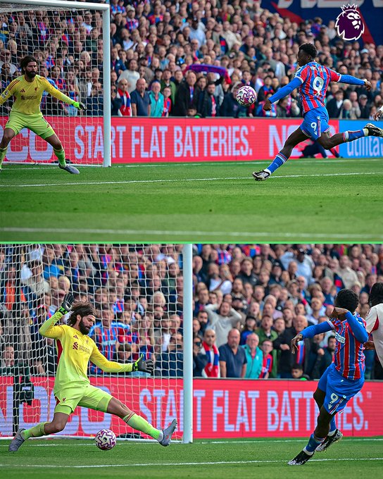 Eddie Nketiah in a blue Crystal Palace uniform, kicking a soccer ball toward a goal. A goalkeeper in a yellow uniform dives to block the shot. The scene occurs on a grassy field with a large crowd in the background. Advertisements with the text "START FLAT BATTERIES" are visible along the sidelines.
