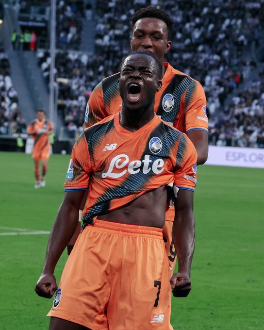 Kamaldeen Sulemana wearing an orange Atalanta B.C. soccer jersey with "Lete" text and the number 7. He is on a grassy field, celebrating with arms raised and an open mouth, with another player in a similar jersey behind him. A crowd is visible in the background.