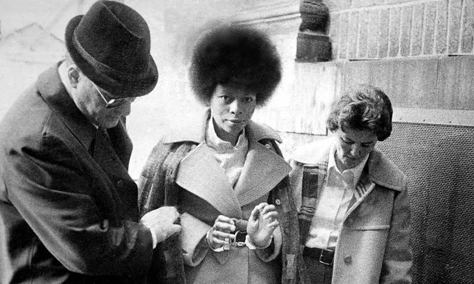 Assata Shakur stands in the center, wearing a coat and holding her hands together, possibly in handcuffs. Two individuals, one wearing a hat and glasses and the other in a coat, stand close to her, appearing to interact. The background shows a textured wall and a metal grate.