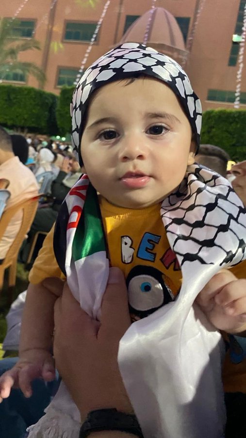 A child wearing a black and white checkered headscarf and holding a Palestinian flag scarf. The child is dressed in a yellow shirt with cartoon designs. An adult hand is visible, holding the child. The background shows a crowd of people and a building with palm trees.