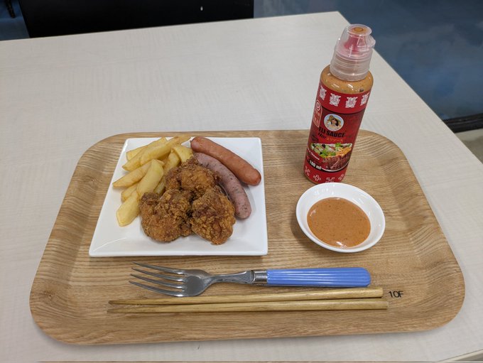 A wooden tray holding a white plate with fried chicken, French fries, and sausages. A small bowl of Eri Hot Sauce is on the tray, with a bottle of Eri Hot Sauce beside it. A fork, knife, and chopsticks are placed on the tray.