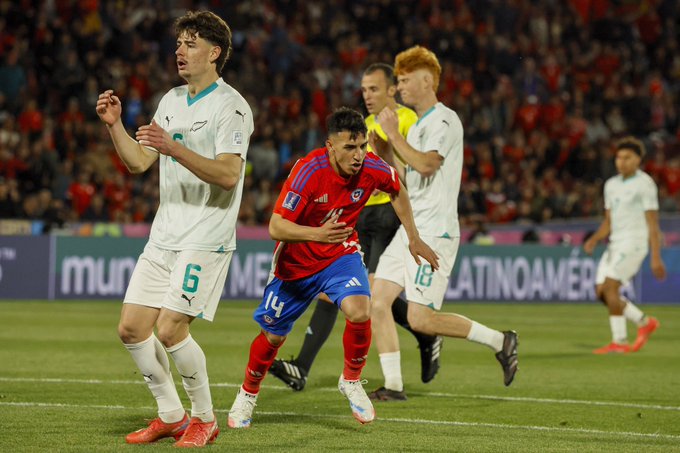 Jugadores de fútbol en acción en un campo. Se ven jugadores con camisetas blancas con el número 6 y otro con camiseta blanca, junto con un jugador con una camiseta roja y azul marcada con el 14. Un árbitro con una camiseta amarilla se encuentra cerca. El fondo muestra una multitud y una pancarta con el texto "Mundial" y "Latinoamérica".