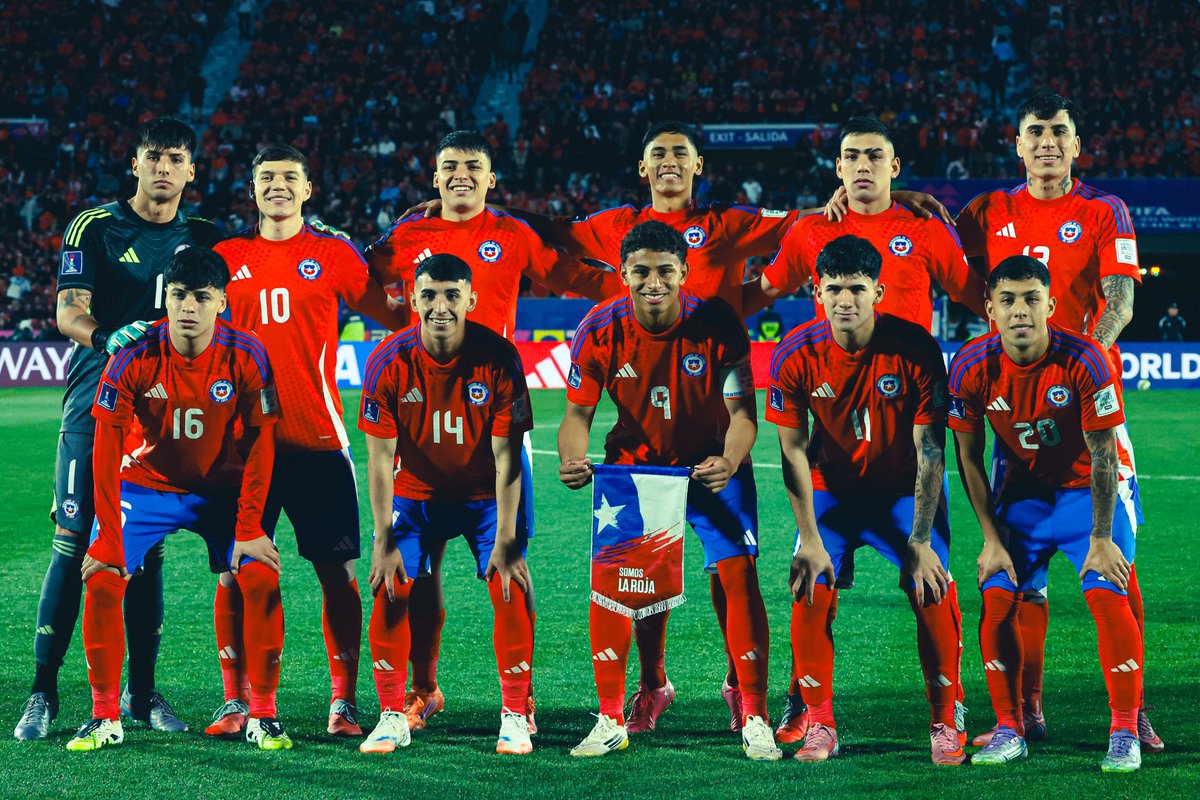 A group of soccer players in red jerseys with blue accents and the number 10 prominently displayed. They wear matching shorts and socks, standing on a grassy field with a stadium crowd in the background. A flag with "Chile" is visible, held by one player in the center.