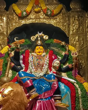 A statue of Kanaka Durgamma, adorned with multiple arms holding various objects, wearing a red and blue sari with gold jewelry and garlands. The statue is seated on a lion, surrounded by colorful flowers and decorations. The backdrop features an ornate golden arch with intricate designs.