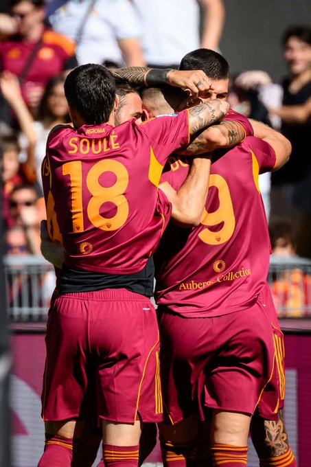 Two soccer players wearing maroon jerseys with yellow accents, one with "SOULE" and the number 18 on the back, the other with the number 9, hugging on a field. Both have visible tattoos on their arms. The crowd and stadium seating are in the background.