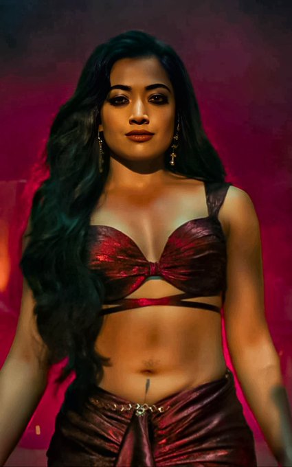 Rashmika Mandanna with long black hair, wearing a red metallic top and matching skirt with a visible tattoo on her navel. She has a serious expression and stands against a red background.