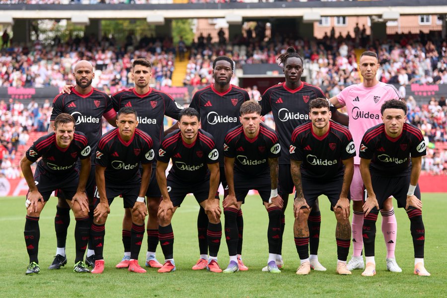Sevilla FC players in black and red uniforms with the Mediaset logo, standing on a soccer field. They are positioned in a line, some crouching and some standing, with a crowd in the background. A goalkeeper in a pink uniform is visible on the right.
