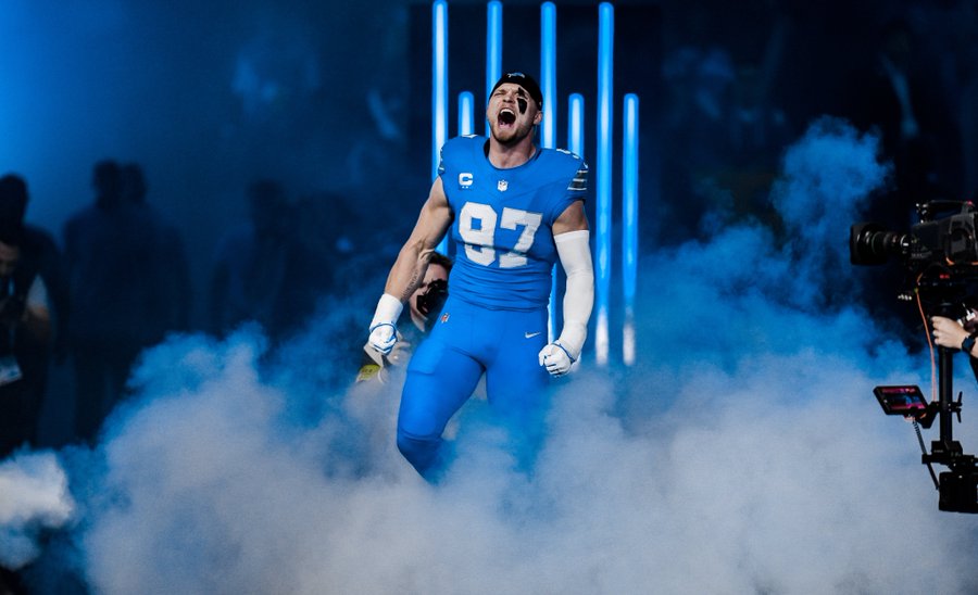 Aidan Hutchinson wearing a blue Detroit Lions jersey with the number 97. He is on a field, surrounded by smoke and blue lights, with a camera visible nearby.