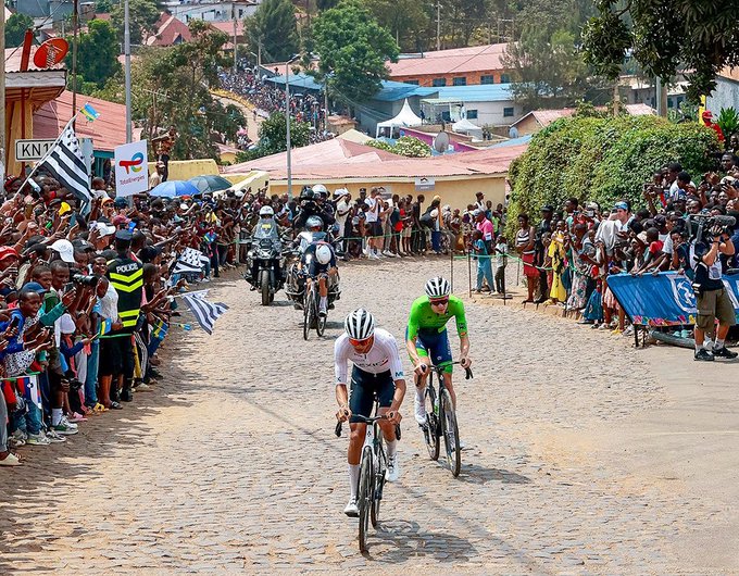 Isaac Del Toro riding a bicycle on a cobblestone road during a race. Another cyclist in a green jersey rides beside him. Crowds of spectators line both sides of the road, cheering. Motorcycles follow closely behind the cyclists. Buildings and greenery are visible in the background.
