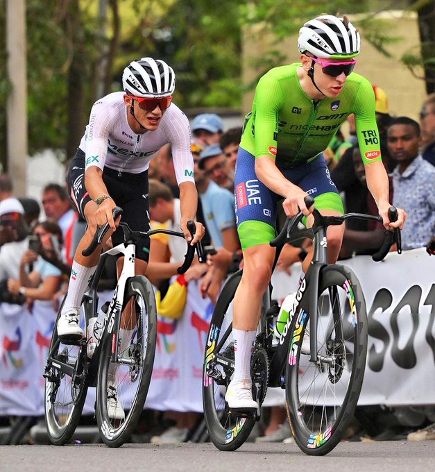 Isaac del Toro and Tadej Pogacar riding bicycles on a road. Both wear helmets and cycling gear, with Isaac in a white and black outfit and Tadej in a green and blue outfit. They are in a competitive race, leaning forward on their bikes with spectators in the background.