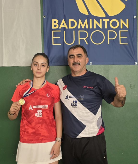 Aleyna Korkut wearing a red sports jersey with "Anadolu" and "SSR" text, holding a gold medal, standing next to a man in a navy and white sports shirt giving a thumbs-up. A blue banner with "BADMINTON EUROPE" in yellow text is visible in the background. Watermarks from Instagram are present.