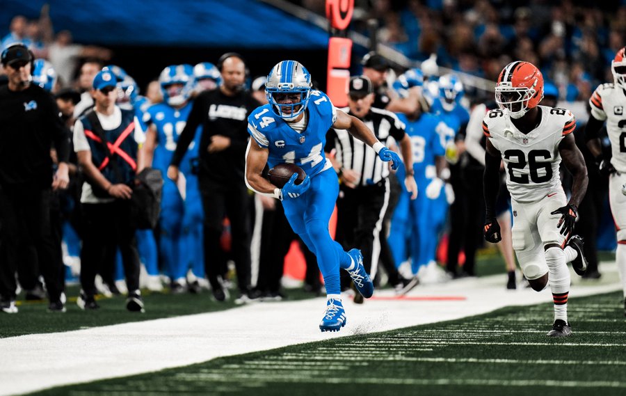 Amon-Ra St. Brown in a blue Detroit Lions uniform running with a football on a field. Calvin Johnson and Leonard Thompson are not visible. Other players in white and brown uniforms, likely Cleveland Browns, are in pursuit. Referees and spectators in blue are visible in the background.